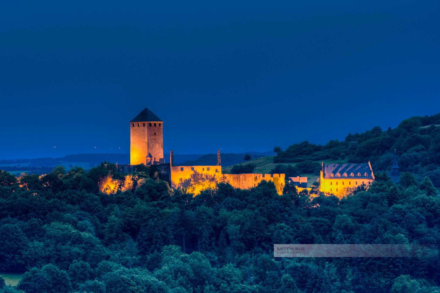 Blaue Stunde an der beleuchteten Burg Lichtenberg. Dunkle Bäume und dunkler Nachthimmel umgeben die Burg